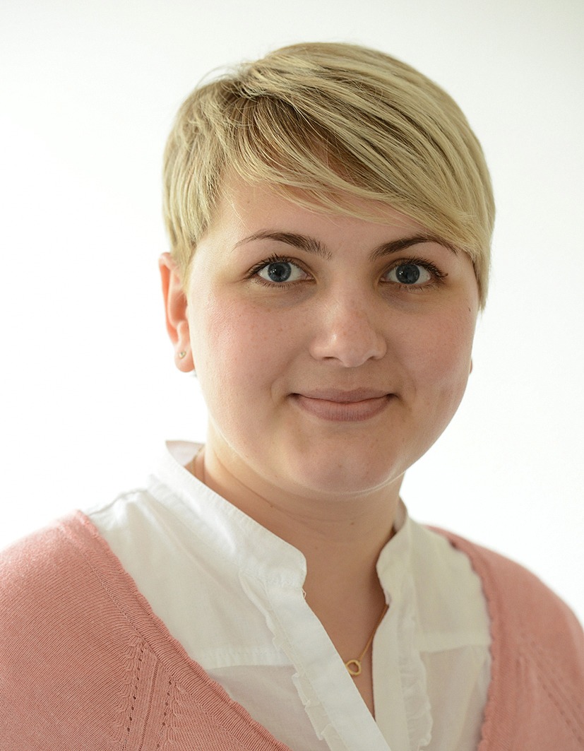 Eine Frau mit kurzen blonden Haaren trägt einen hellrosa Pullover über einem weißen Hemd mit Kragen. Sie lächelt sanft und schaut in die Kamera vor einem schlichten, hellen Hintergrund.