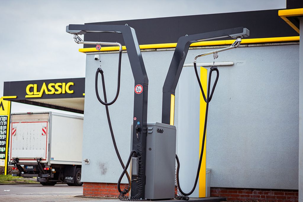 Eine moderne Staubsauger-Selbstbedienungsstation mit zwei hohen Armen und Schläuchen steht vor einer CLASSIC-Tankstelle; im Hintergrund sind Kraftstoffpreise und ein weißer Lieferwagen zu sehen.