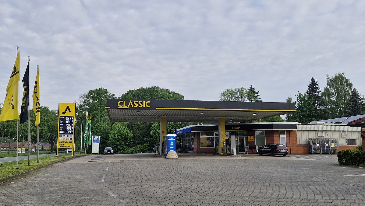 Eine kleine CLASSIC-Tankstelle mit einem Lebensmittelgeschäft, gelber und schwarzer Beschilderung, mehreren Zapfsäulen, einem schwarzen Auto und hohen gelben Fahnen, umgeben von Bäumen und einem teilweise bewölkten Himmel.