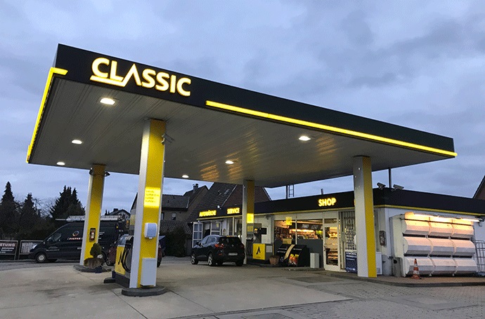 Eine moderne Tankstelle mit gelber und schwarzer Beschilderung mit der Aufschrift CLASSIC. Autos parken neben den Zapfsäulen und dem Laden, der Himmel ist wolkenverhangen und im Hintergrund sind mehrere Gebäude zu sehen.