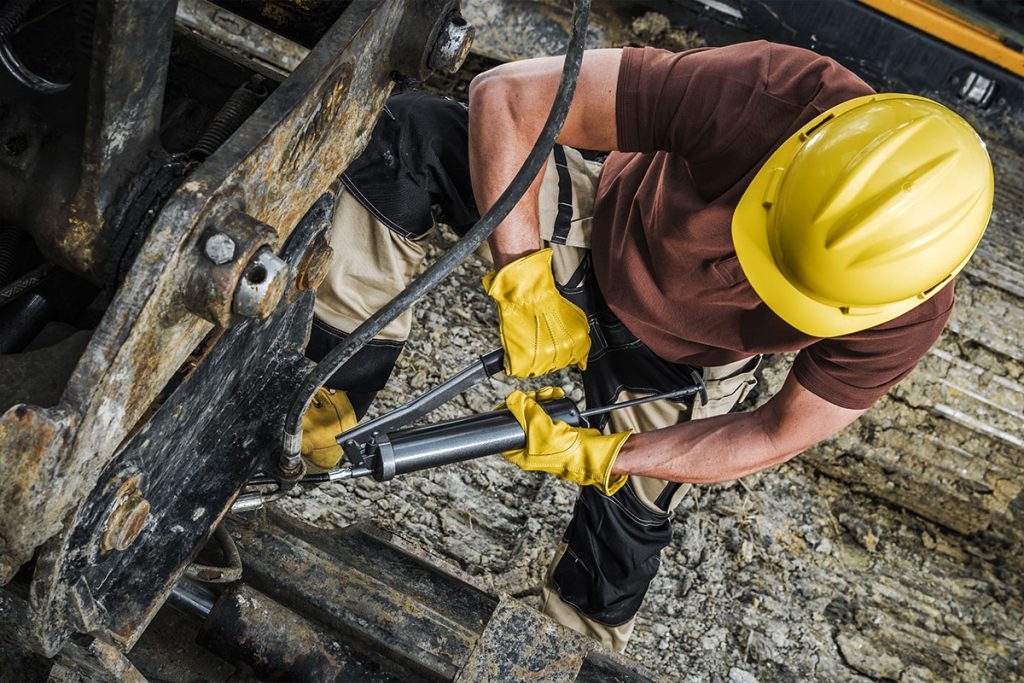 Ein Bauarbeiter, der einen gelben Schutzhelm, Handschuhe und Schutzkleidung trägt, verwendet eine Fettpresse, um schwere Maschinen im Freien auf einer Baustelle mit Schmutz und Steinen zu warten.