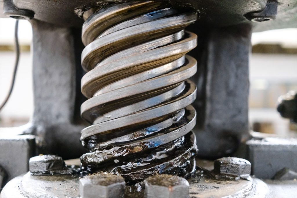 Nahaufnahme eines mit dunklem Fett beschichteten Metallzahnrads oder einer Schnecke, die Teil einer Industriemaschine ist. Die Oberfläche weist Verschleiß- und Gebrauchsspuren auf, an der Basis sind Schrauben sichtbar.