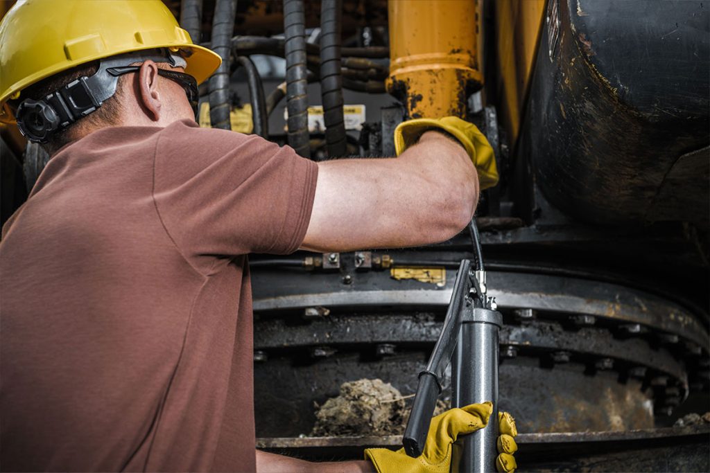 Ein Arbeiter mit einem gelben Schutzhelm und Handschuhen schmiert mit einer Fettpresse Maschinenteile an einem großen Industriefahrzeug.