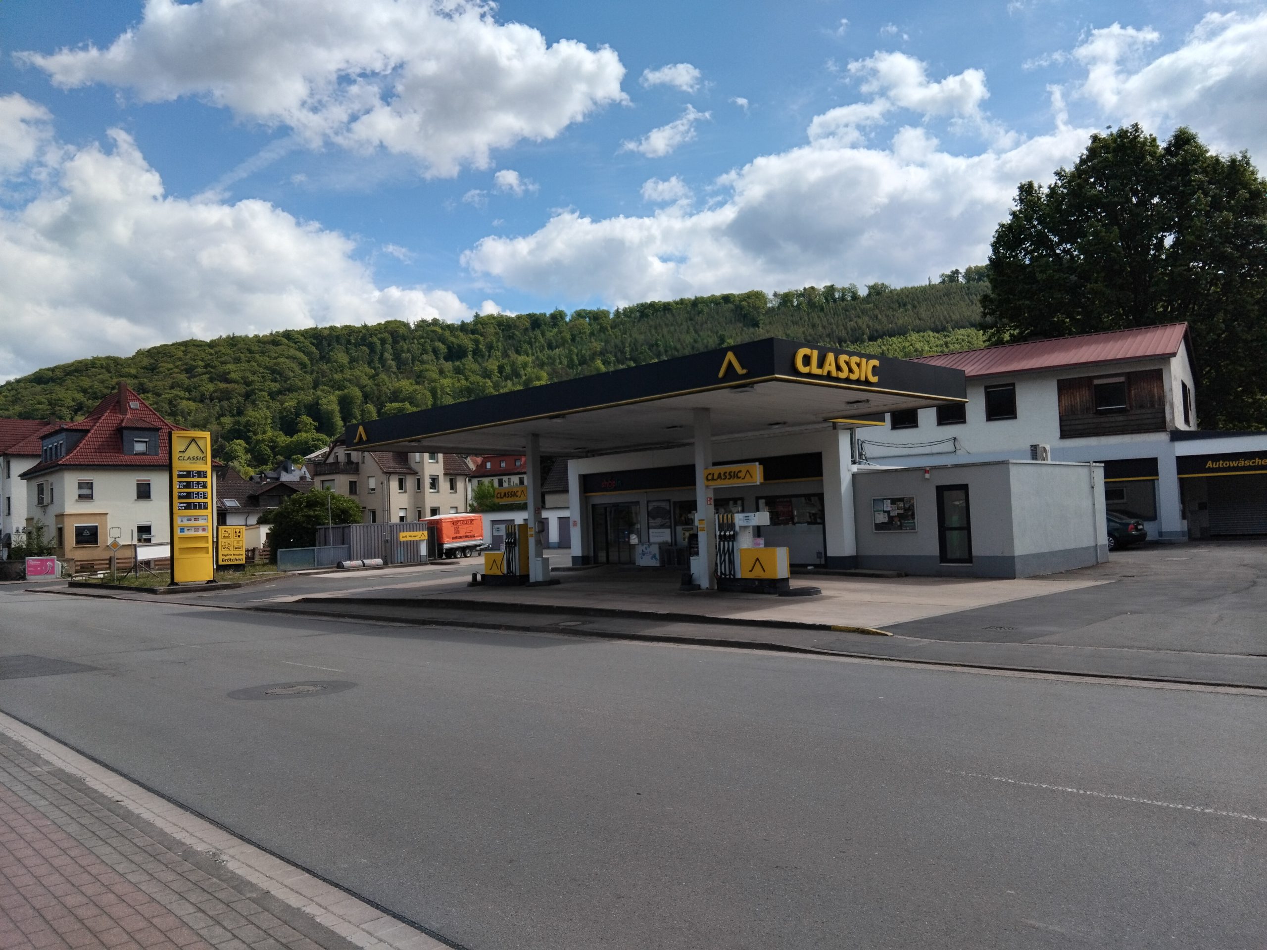 Eine CLASSIC-Tankstelle mit einem gelb-schwarzen Schild liegt an einer ruhigen Straße in einer Kleinstadt, umgeben von Häusern und grünen, baumbewachsenen Hügeln unter einem teilweise bewölkten Himmel.