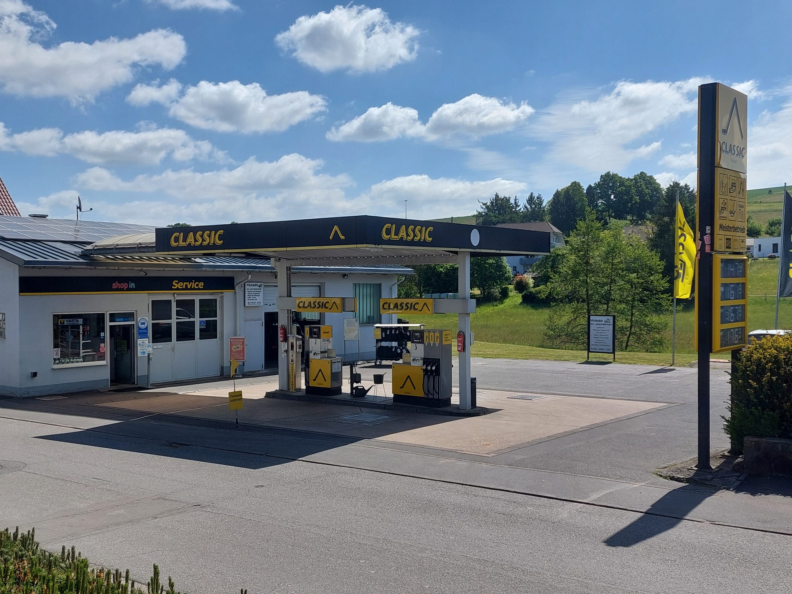 Eine kleine Tankstelle der Marke Classic mit zwei gelb-schwarzen Zapfsäulen, einer Werkstatt und einem großen Schild am Straßenrand in einer ländlichen Gegend mit grünem Gras und Bäumen unter einem blauen Himmel mit Wolken.