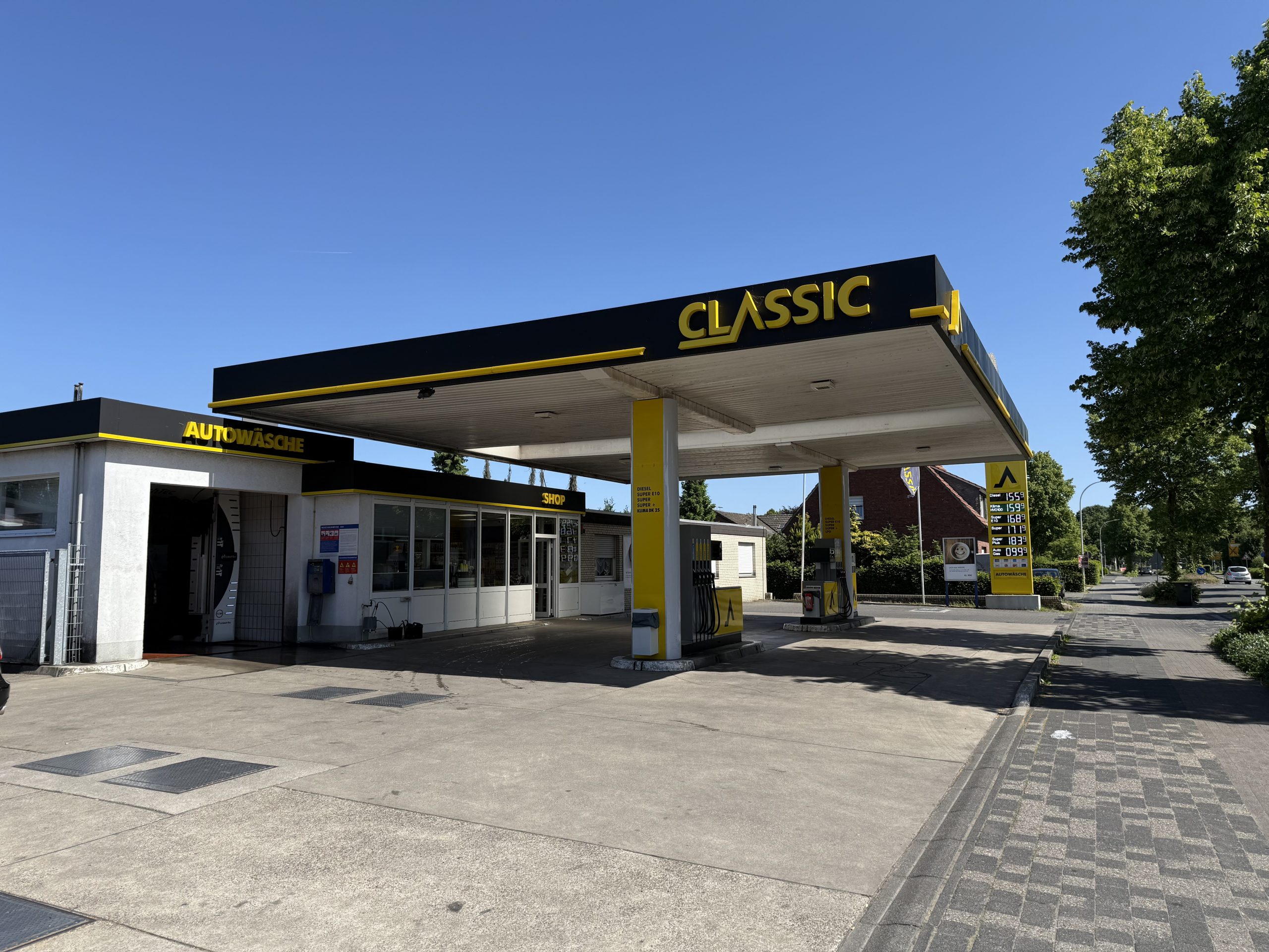 Eine CLASSIC-Tankstelle mit gelbem und schwarzem Branding, mehreren Zapfsäulen und einem kleinen Nebengebäude mit der Aufschrift "AUTOWÄSCHE" an einem sonnigen Tag mit blauem Himmel und einigen Bäumen im Hintergrund.