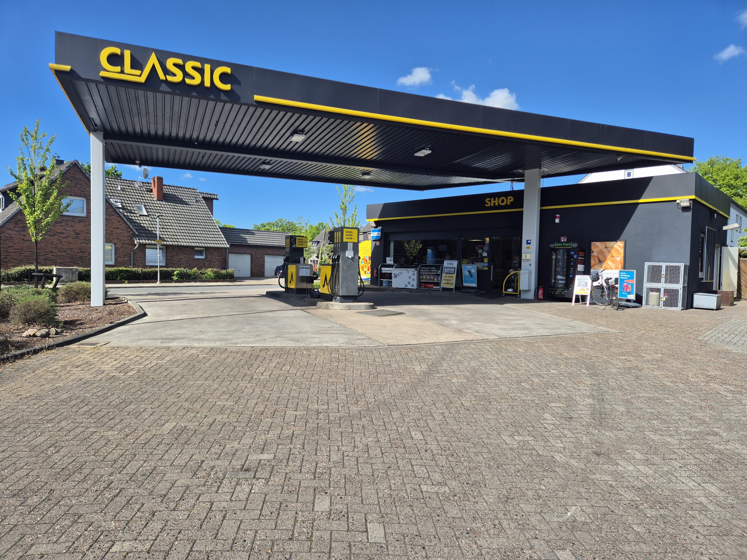 Eine kleine Tankstelle namens CLASSIC mit gelbem und schwarzem Branding, eine Zapfsäule und ein Ladengebäude. Der Himmel ist klar und blau, und im Hintergrund sind Häuser und Bäume zu sehen.