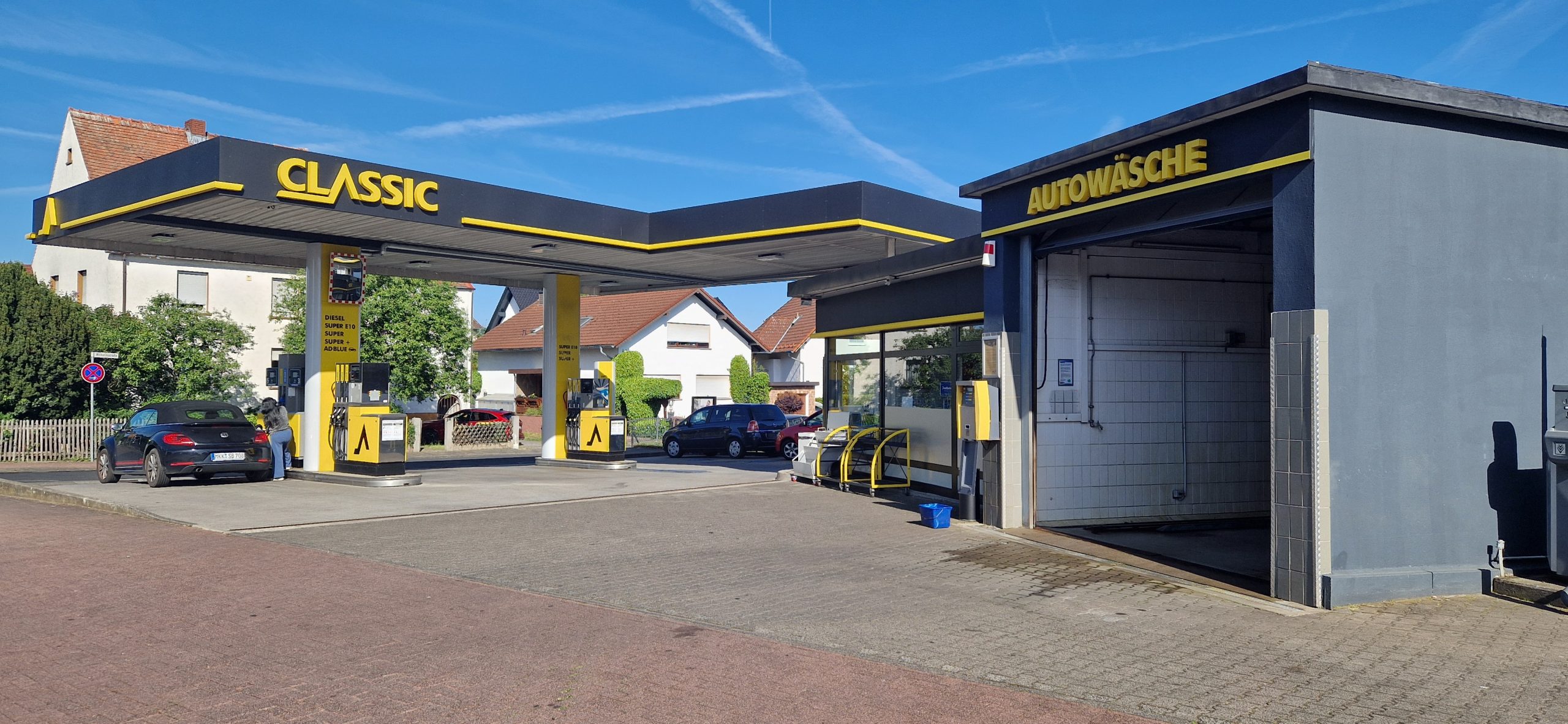 Tankstelle mit gelb-schwarzem CLASSIC-Schild, zwei tankende Autos und rechts ein kleines Gebäude mit der Aufschrift AUTOWÄSCHE für die Autowäsche. Im Hintergrund sind Wohnhäuser und Bäume unter einem klaren Himmel zu sehen.