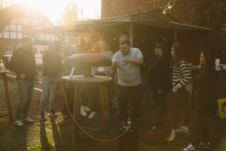 Eine Gruppe von Menschen steht draußen um einen holzbefeuerten Pizzaofen herum, lächelt und lacht im Sonnenlicht. In der Nähe liegt ein Hund im Gras, und über ihm hängen Lichterketten. Die Szene ist warm und zwanglos, mit einer entspannten, sozialen Atmosphäre.