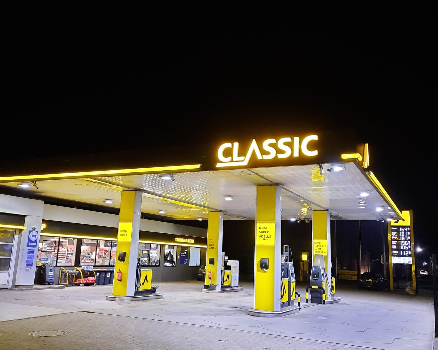 Eine hell erleuchtete CLASSIC-Tankstelle bei Nacht, mit gelber Beschilderung und Säulen, Zapfsäulen unter dem Vordach und einem Lebensmittelgeschäft im Hintergrund. Die Umgebung ist dunkel.